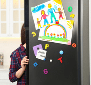A woman opening a refrigerator that has a child's artwork posted on the door. 