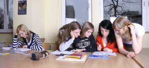 A teenage girl is sitting apart from the group reading a book while four other teenage girls are gather around each other at the end of the table.