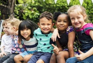 six children of diverse ethnicities are sitting arm in arm smiling for the camera
