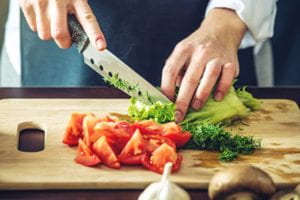 Close up of two hands using a large knife to cut vegetables.