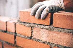 A gloved hand places a brick in a wall under construction.