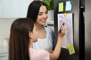Mother and daughter writing on a chore chart that is posted on the refrigerator.