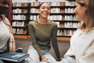Three smiling young people sitting on the floor of a library with a large bookcase behind them.