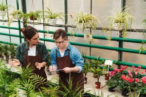 A young man takes direction from an adult woman as they work together in a greenhouse.