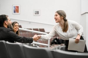 Student with papers in her hand leaning over a seat in an auditorium to shake the hand of a bearded man while another woman looks on from the background.