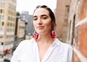 Head shot of young woman with long dangling red earrings, looking confidently at the camera with a cityscape behind her. 