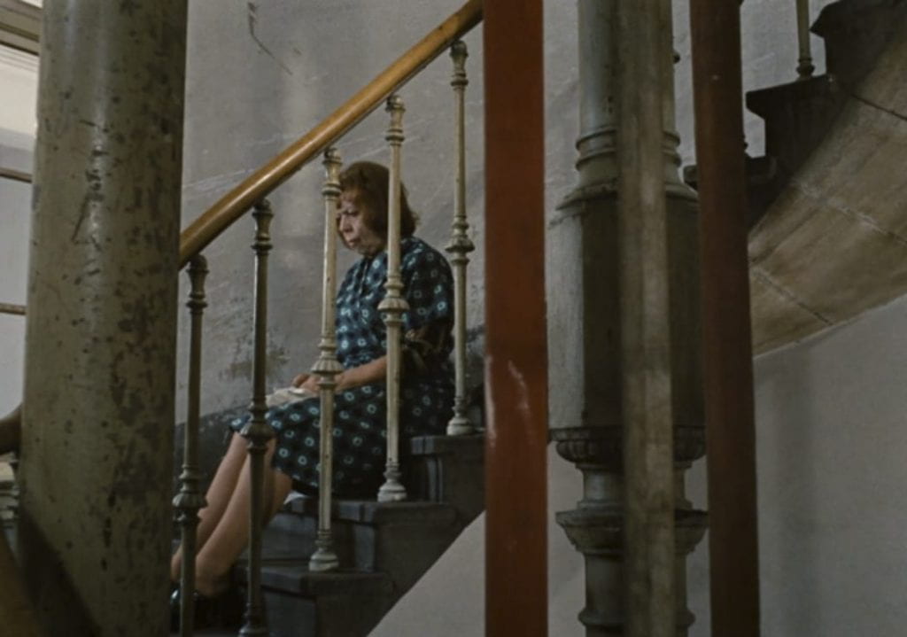 Emmi is now alone, which is visually marked by her framing and fragmentation between the stair's railing.