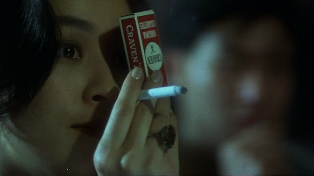 A woman holds up a pack of cigarettes to cover one of her eyes while smoking