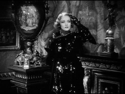 Marlene Dietrich in a shimmering gown stands in an ornately decorated room