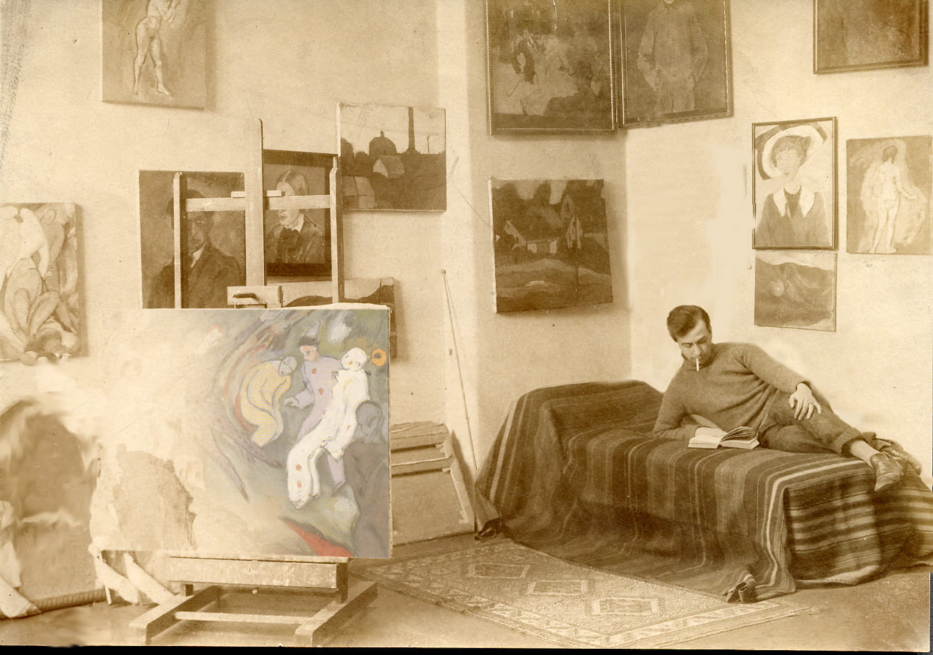 A sepia-toned image of a man smoking and reading on his bed in a room surrounded by paintings