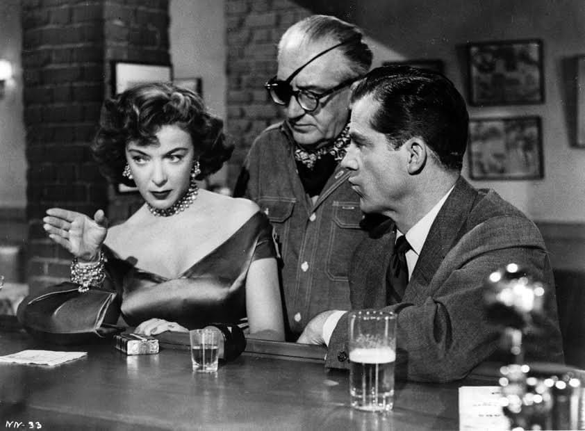 A man with an eyepatch stands behind a second man and a woman who are seated at a bar