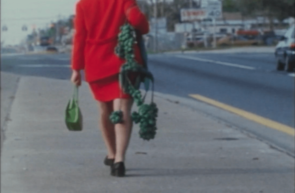 A woman in a red skirt-suit walks down the sidewalk, carrying heavy, metal protrusions coming out of the back of her dress like a tail.