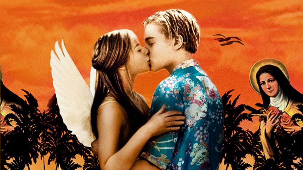 A teenage girl wearing angel wings kisses a teenage boy in a Hawaiian shirt against an orange sky