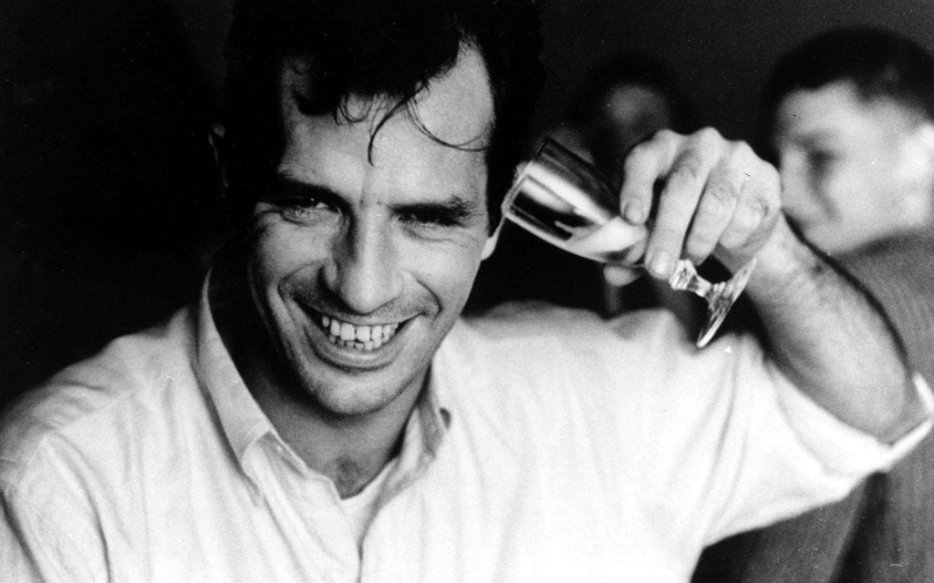 Black and white image of a man smiling and holding a glass of liquor up to his ear
