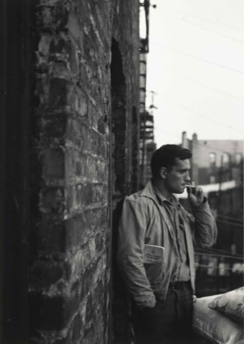 A man stands in an alleyway with a book in his jacket and smoking a cigarette