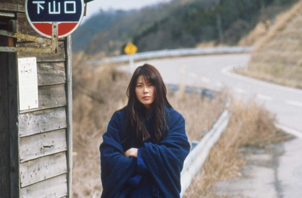 A woman wrapped in a blanket stands by the side of the road