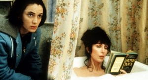 Winona Ryder looks offscreen while Cher reads a book in a bubble bath