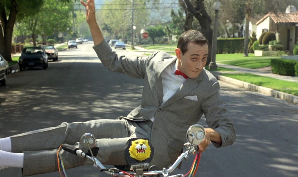 Pee-wee shows off on his back, riding with his legs out to the side and with only one hand on the handlebars.
