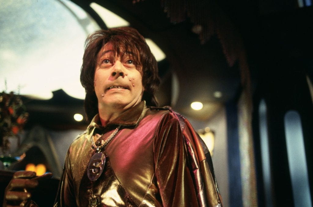Paul Reubens as The Spleen wears a shiny gold and maroon cape. He looks up, his face tensed and his zits prominent.