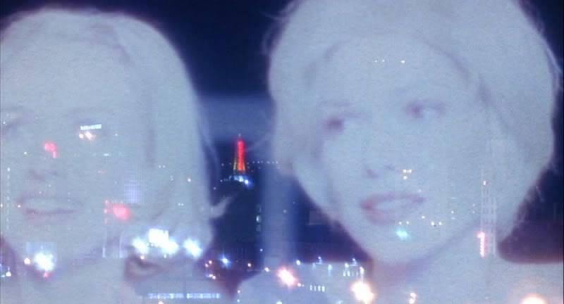 The faces of two smiling blonde women are projected against a background of city lights at night