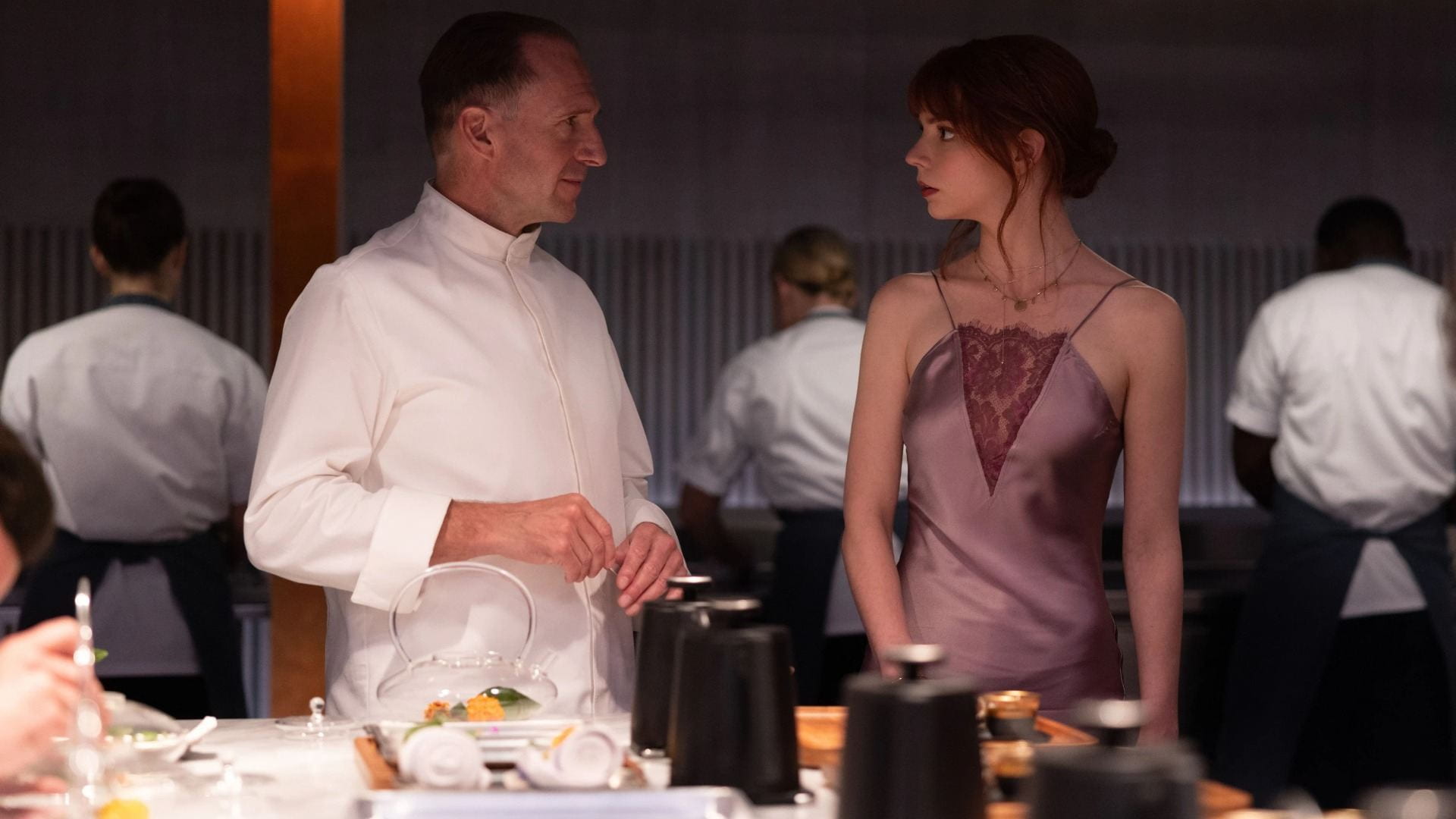 A man in a chef's uniform and a woman in a sleek dress look at each other in a busy kitchen