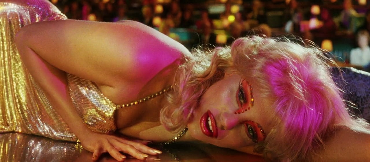 Close-up of a blonde woman with elaborate eye make-up and a gold dress laying on a stage