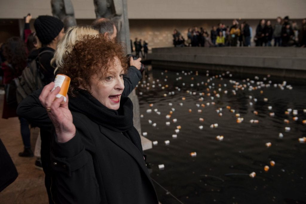 Nan Goldin holds a prescription medication bottle and points to a floor littered with more bottles while spectators watch