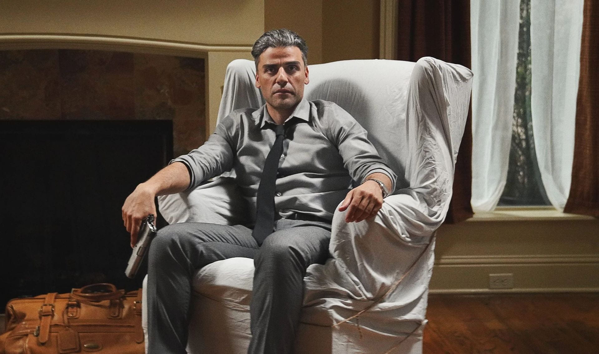 Oscar Isaac holds a gun and sits in a chair that's covered in a white cloth