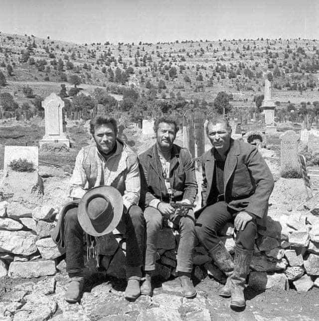 Clint Eastwood, Eli Wallach, and Lee Van Cleef sit on a rock wall in a cemetery