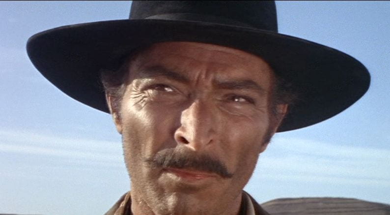 Close-up of a squinting, mustachioed man in a black cowboy hat with a blue sky behind him