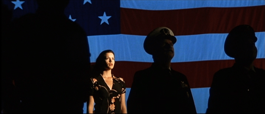 Grace (Liv Tyler) standing in front of a U.S. flag watching the launch. She is lit with warm light while the soldiers in the foreground are silhouetted