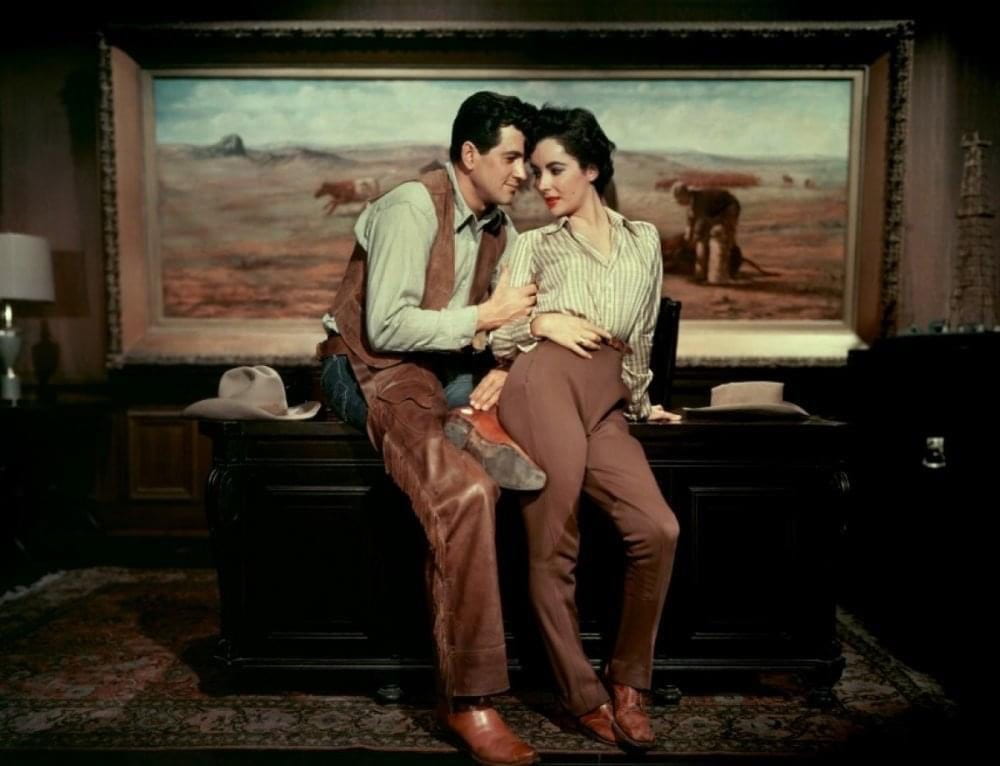 Rock Hudson sidles up to Elizabeth Taylor as they sit on a desk in a wood-clad office