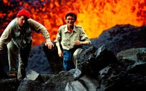 A man and woman smile at the camera while a volcano erupts lava behind them