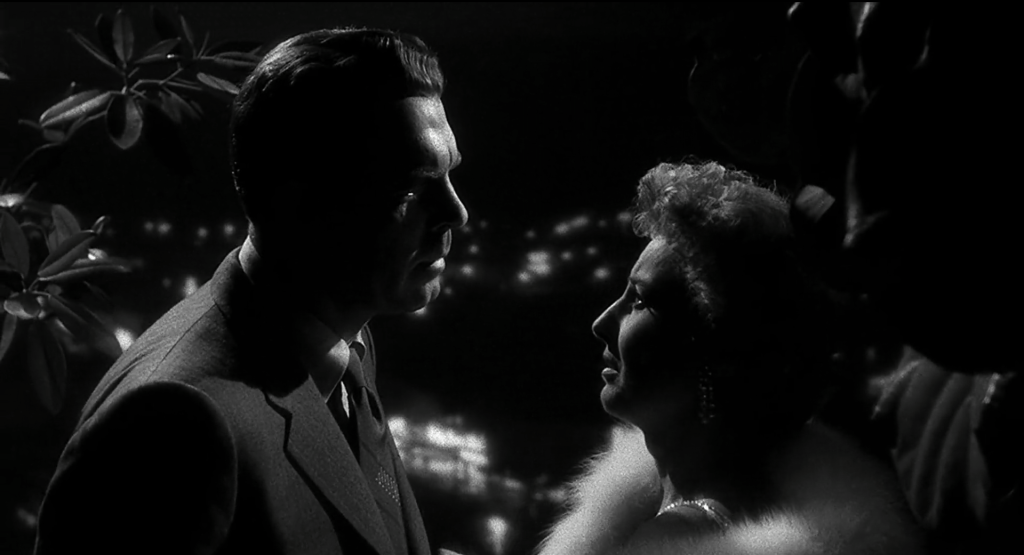 A two-shot of a man in a suit and a woman in a fur coat as they look at each other with city lights in the background