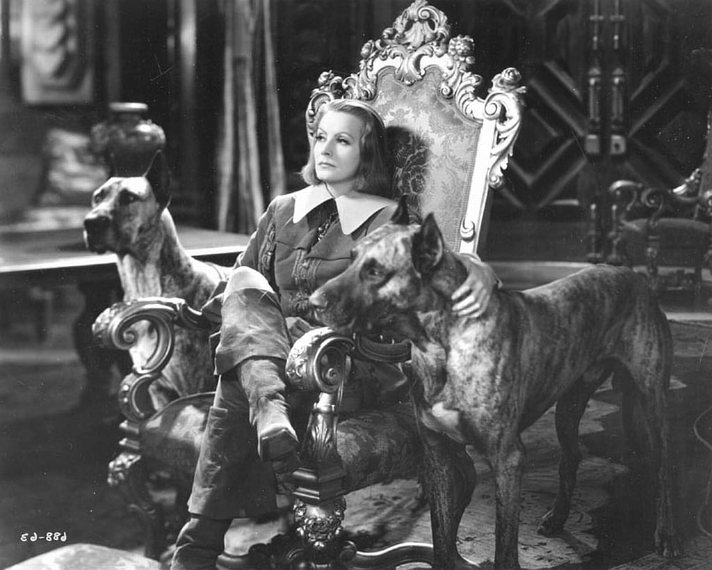 Greta Garbo sits with her legs crossed on a throne with two giant dogs on each side