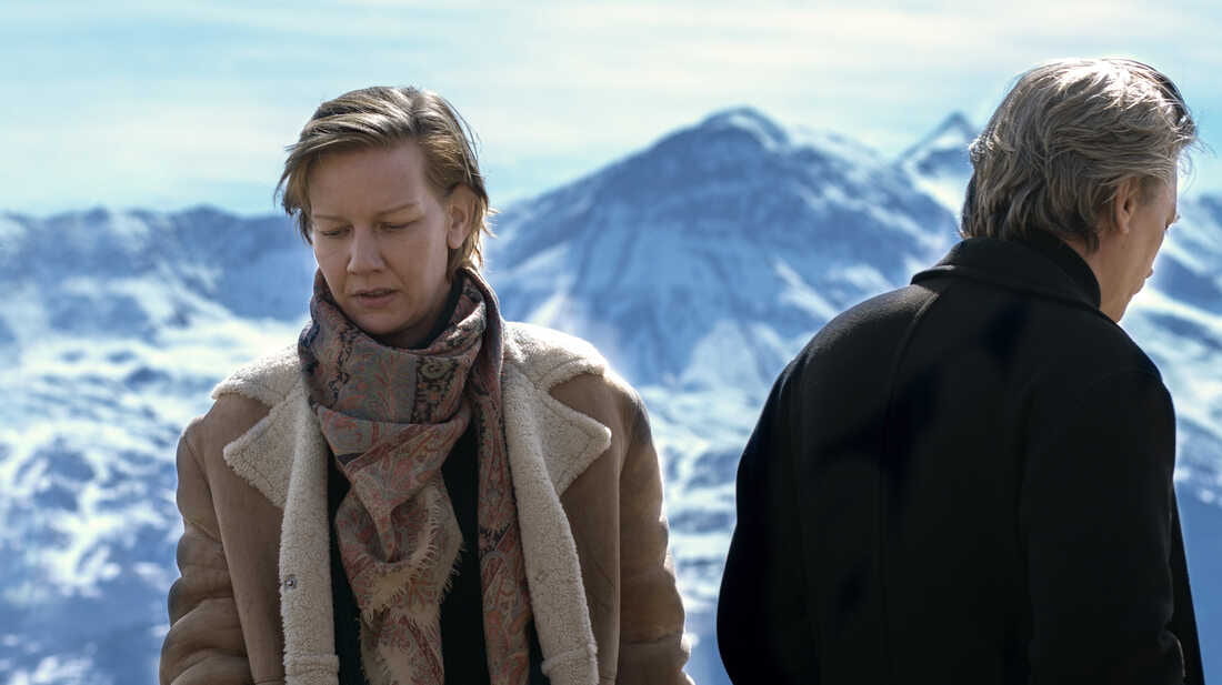 A woman and man stand outside with a mountain behind them, her facing the camera and him with his back to the camera