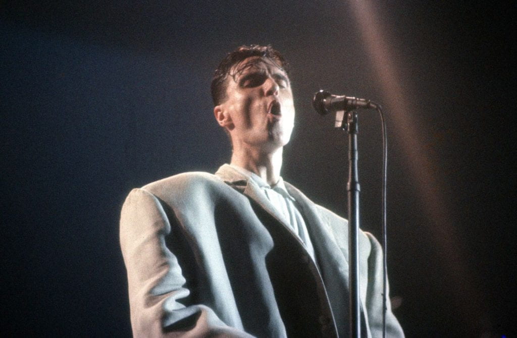 David Byrne sings into a microphone while wearing his big suit