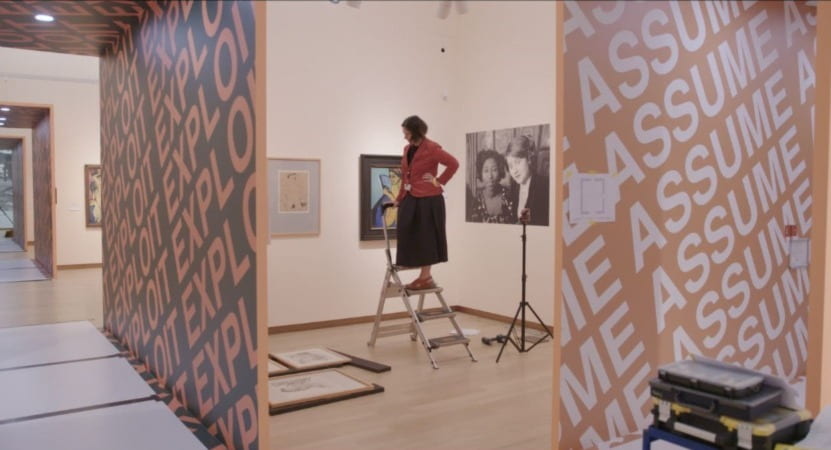 A woman stands on a stepladder in a museum gallery while looking down at artwork that's on the floor