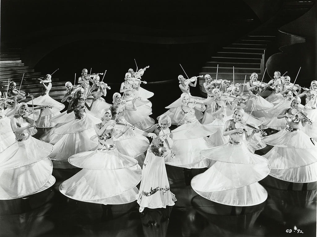 Chorus girls in hooped skirts dance on a stage holding violins