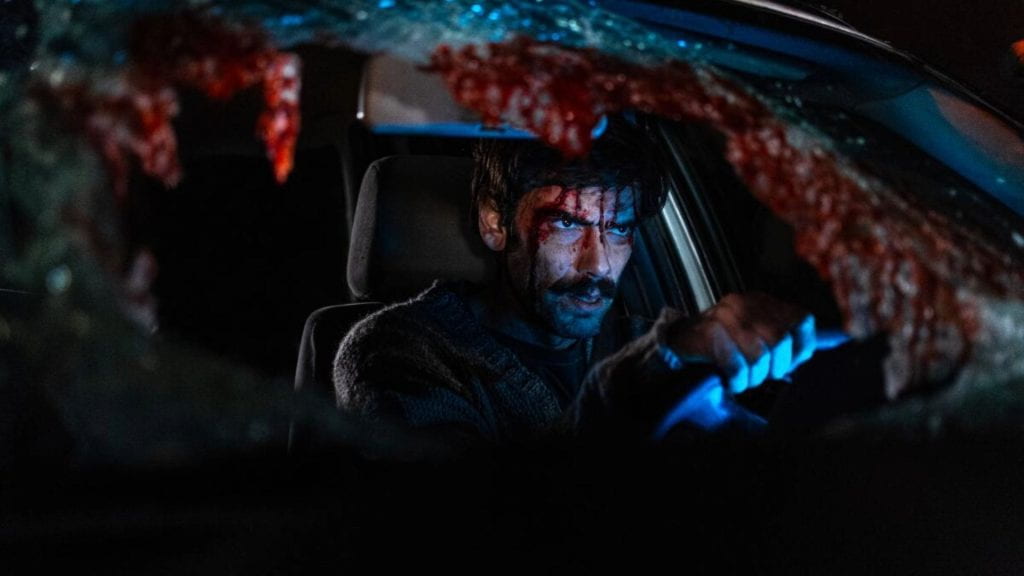 A man with a bloodied face sits in a car with a shattered windshield that also has blood on it
