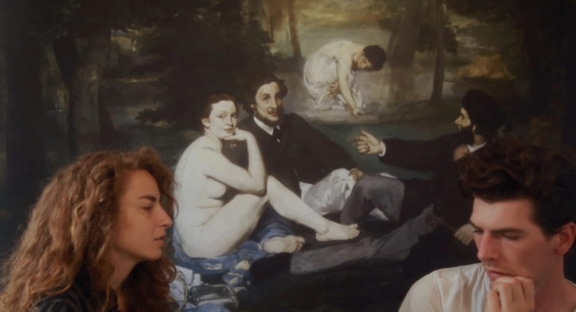 A woman and man sit in front of a painting of people who are also sitting