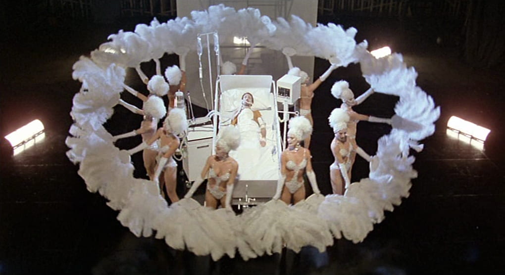 A group of showgirls with feathery fans surround Joe in a hospital bed