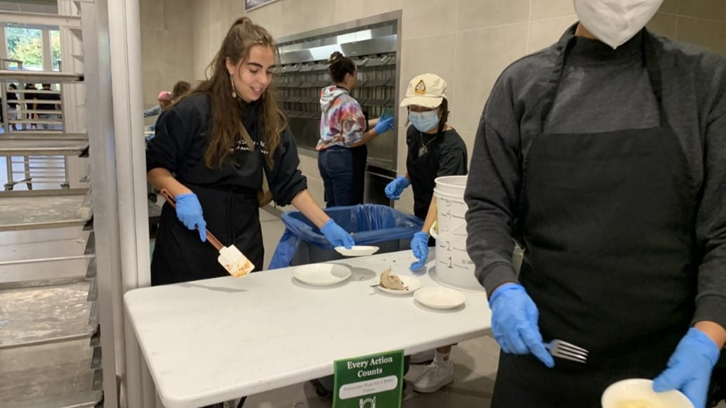Two students receive plates to be scraped. One student is wearing a ponytail and the other is wearing a hat, two braids, and a surgical mask. Both students are wearing blue latex gloves and black aprons.