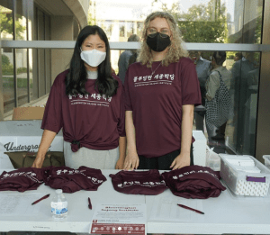 Two graduate students wearing tshirts with Korean writing, standing behind a table with folded tshirts that match theirs.