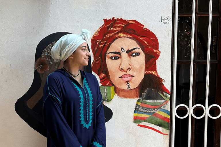 Sofia standing next to a wall with a painting of a woman in a red head covering