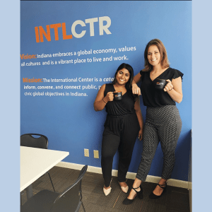 Neelam Patel and Kathryn Petersen pose by a wall reading "INTL CTR Vision: Indiana embraces a global economy, values all cultures and is a vibrant place to live and work. Mission: The International Center is a catalyst to infomr, convene, and connect public, private, and civic global objectives in Indiana.