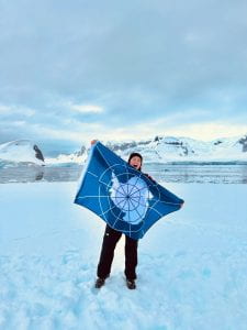 Woman standing on snowy ground in Antarctica, holding blue flag with Antarctica outlined on it in white. Water and glaciers are seen in the background