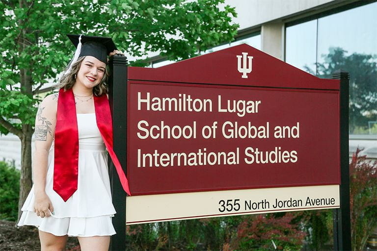 Mackenzie Knight, wearing a graduation cap and red graduation sash, leans against an exterior sign reading "Hamilton Lugar School for Global and International Studies."