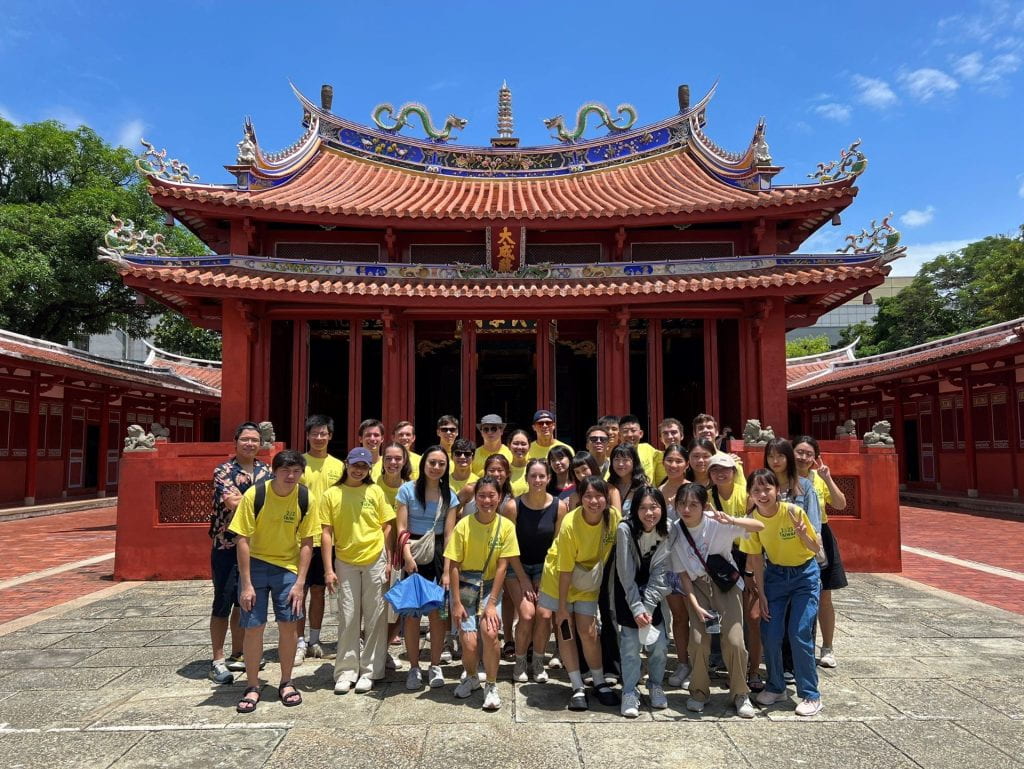 A group of students in yellow shirts pose in front of a temple with an ornate red roof.