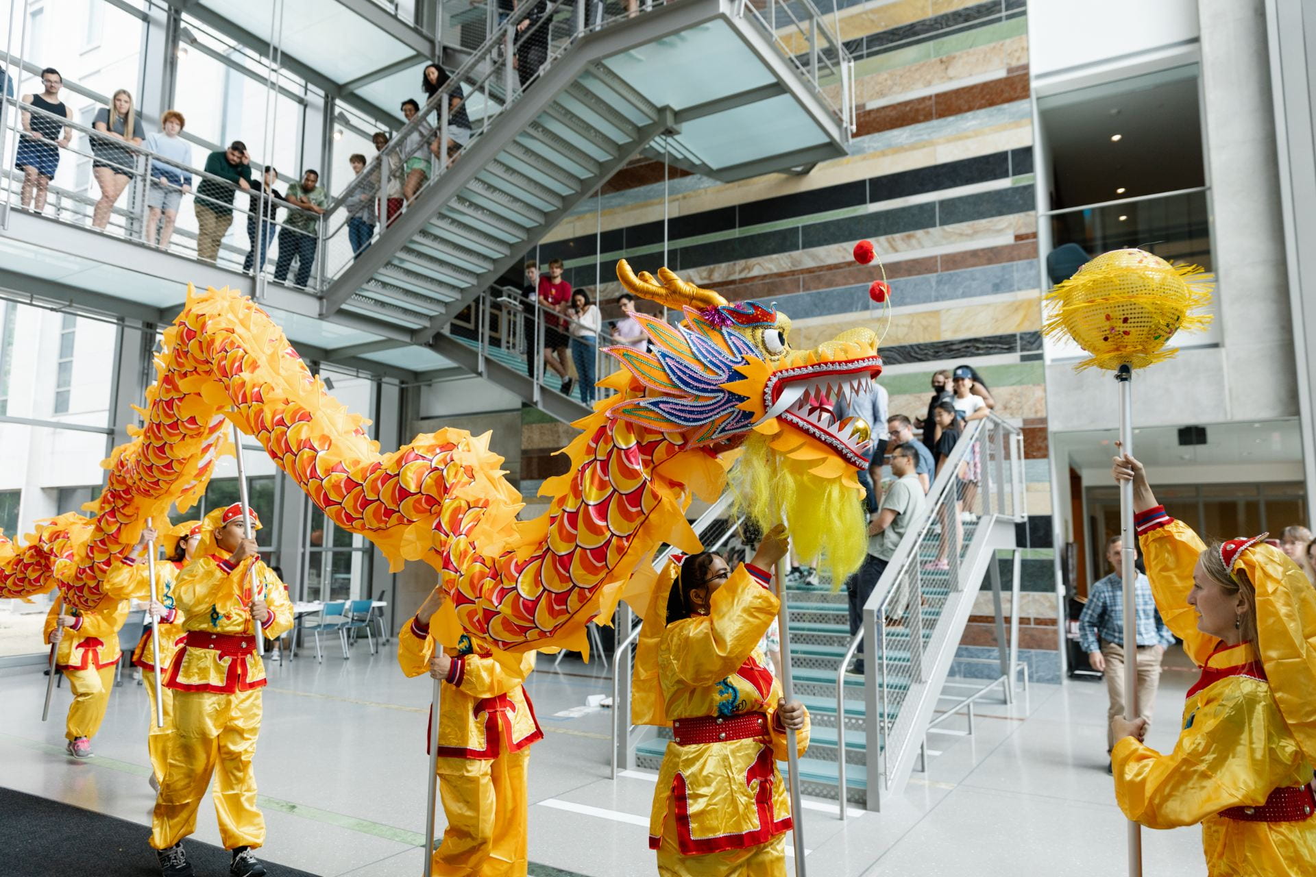 In the Hamilton Lugar School atrium, a colorful Gold, orange, and red paper dragon is held aloft on poles by five students in gold and red matching outfits. Many people line the stairs and walkway above the atrium, watching the display.